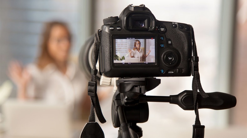 Close up image of camera on tripod with smiling woman on back screen and blurred scene on background. Recording video on modern DSLR camera. Personal videoblog social network concept