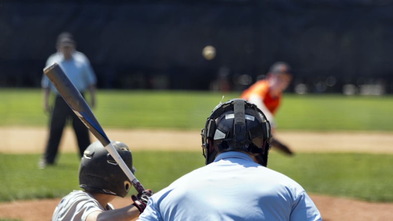 view of an umpire standing behind a batter as pitch comes in during a youth baseball game