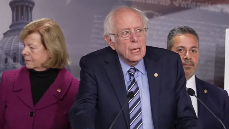 Sen. Bernier Sanders speakers at a press conference. Sen. Tammy Baldwin and Ben Ray Lujan appear behind him. 
