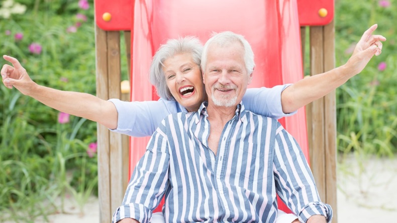 an older man and woman happily slide down a playground slide 