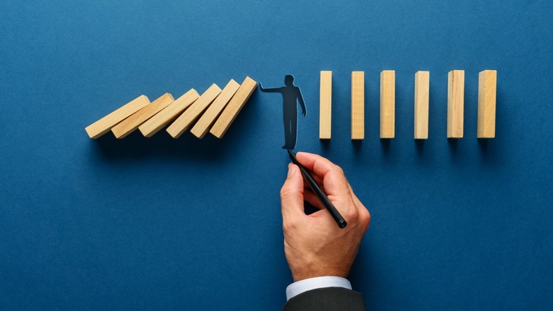 Wide view image of male hand drawing silhouette of a man making a stop gesture to prevent wooden dominos from collapsing. Over navy blue background.